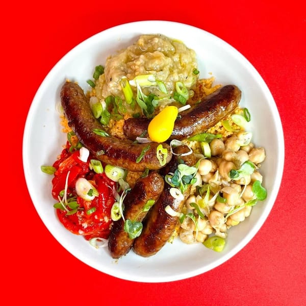 A white bowl filled with sausages, couscous, chickpeas, red peppers, and green onions.