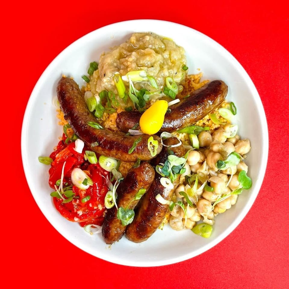A white bowl filled with sausages, couscous, chickpeas, red peppers, and green onions.