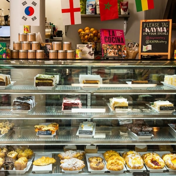 A glass display case filled with various cakes, pastries, and muffins at Kayma restaurant.