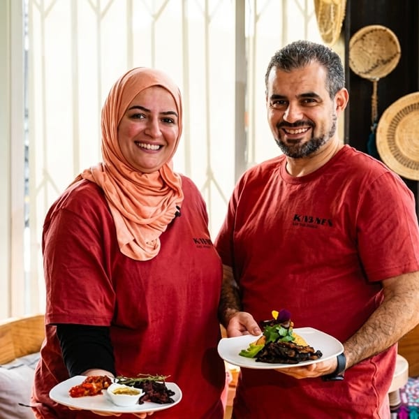 Two smiling chefs in red shirts hold plates of Middle Eastern food in a restaurant.