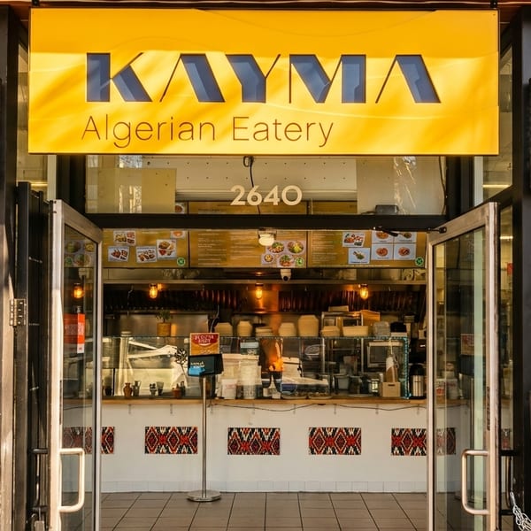 Exterior of Kayma Algerian Eatery with open glass doors, a bright yellow sign, and visible kitchen.