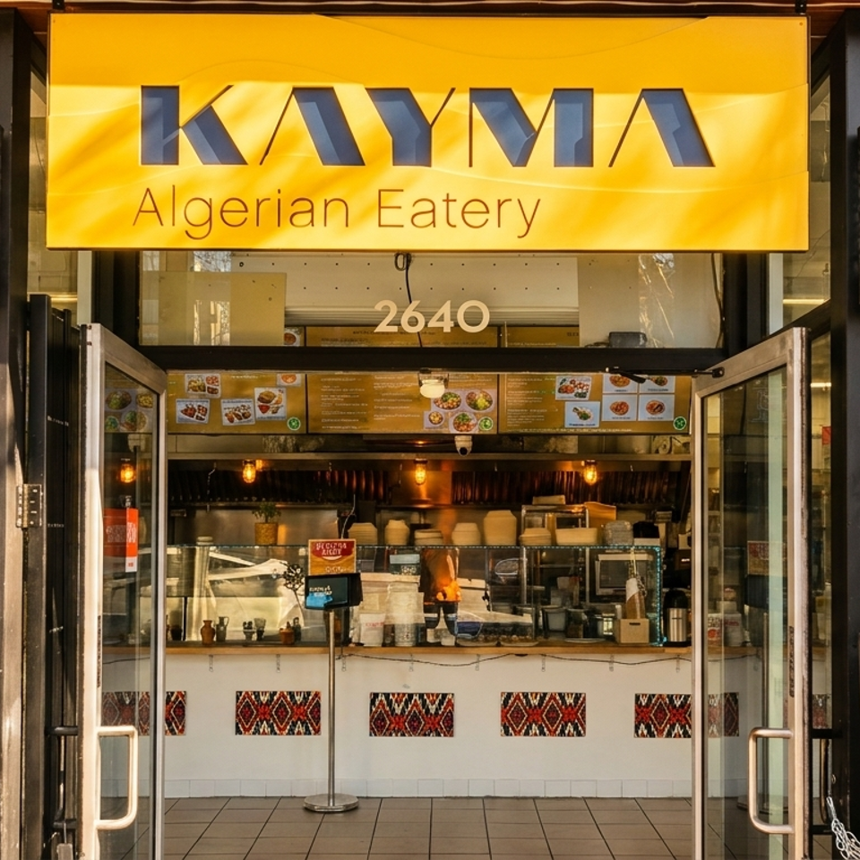 Exterior of Kayma Algerian Eatery with open glass doors, a bright yellow sign, and visible kitchen.